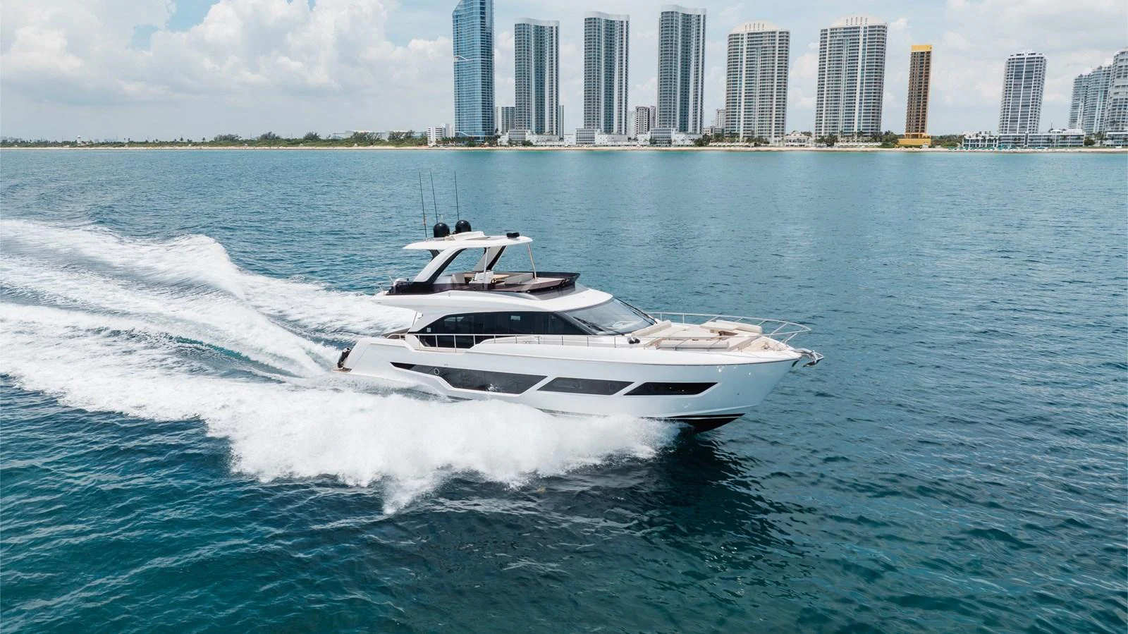 A sleek yacht cruises swiftly on the water with a city skyline in the background. aboard KATRIN I Yacht for Sale