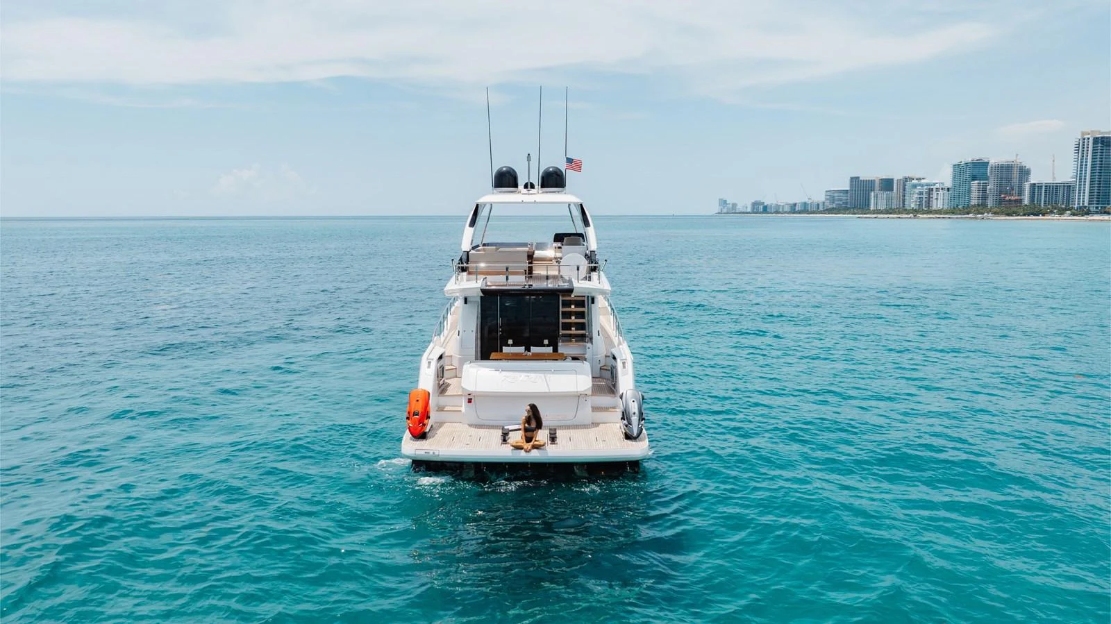 A sleek yacht is seen from the rear, cruising in calm, clear waters with a cityscape in the background. aboard KATRIN I Yacht for Sale