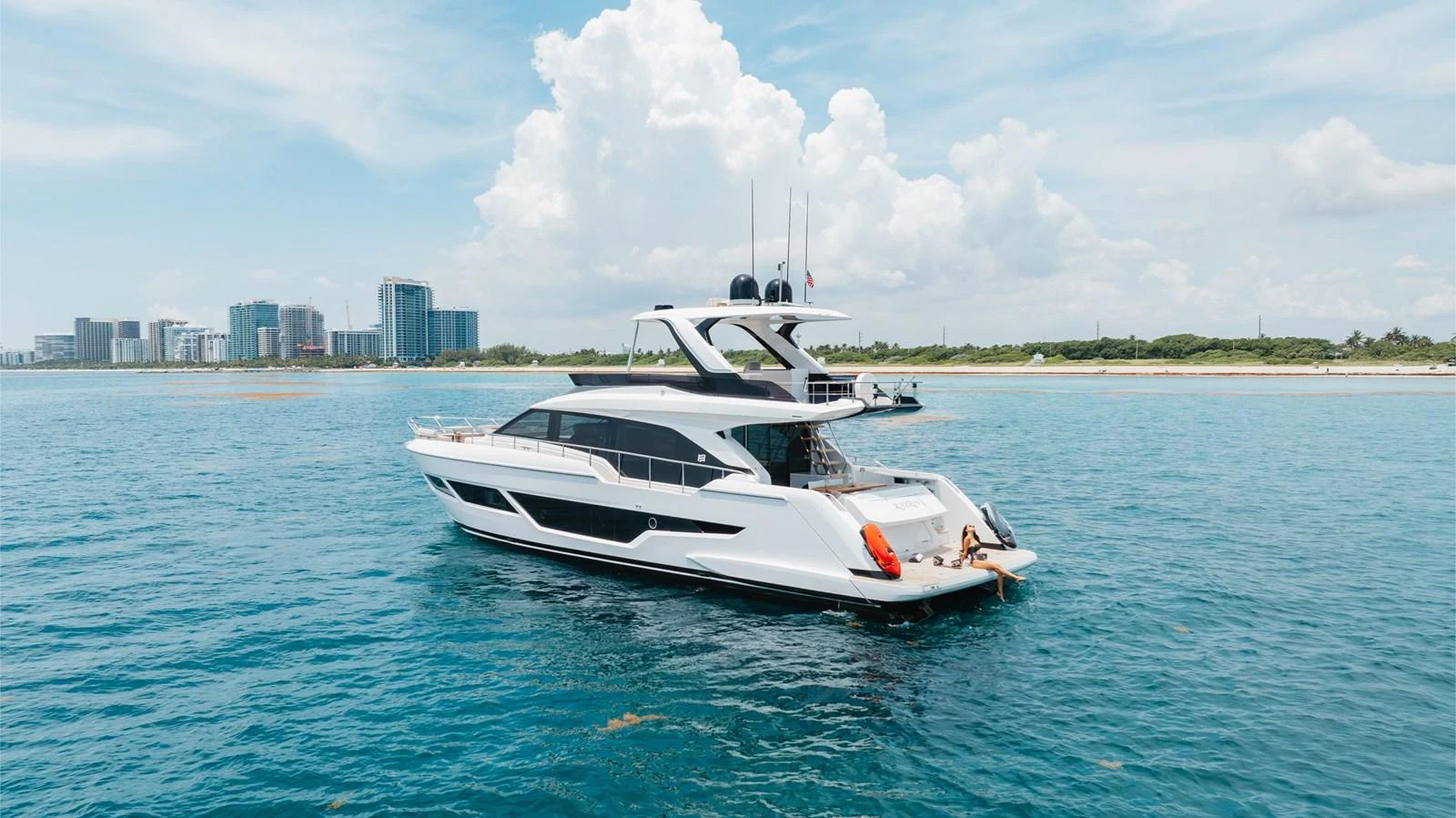 A sleek and modern yacht cruises gracefully on calm blue waters with a cityscape in the background. aboard KATRIN I Yacht for Sale
