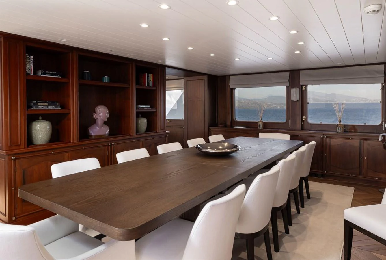 The yacht's dining area features a long wooden table surrounded by white chairs, with large windows offering a scenic view. aboard PANACHE Yacht for Sale