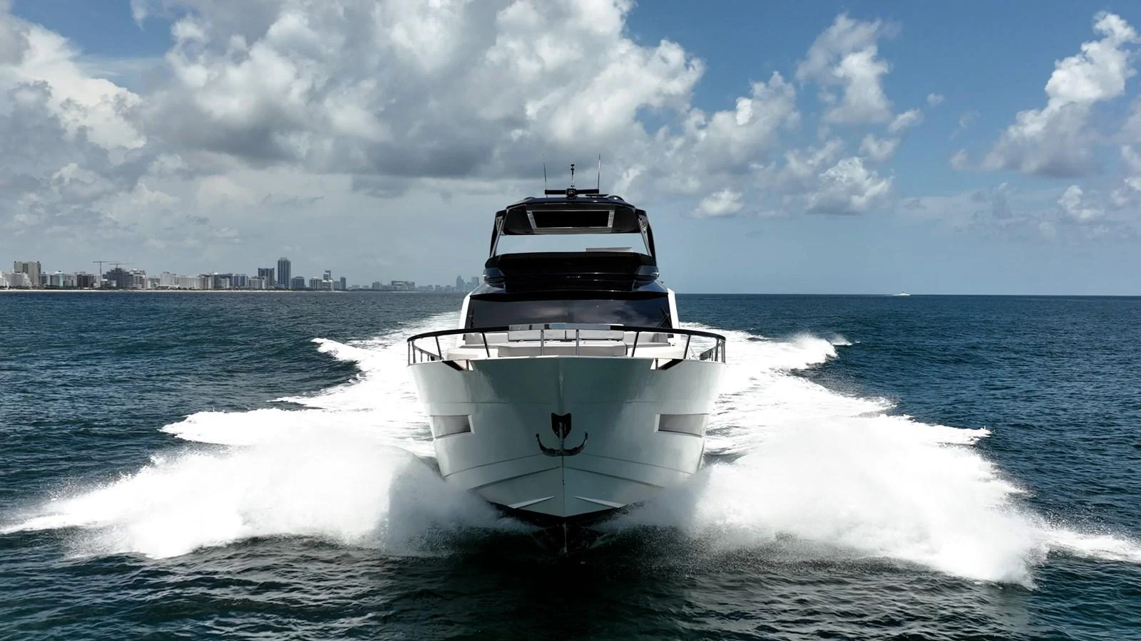 A sleek yacht cuts through the water, showcasing its powerful presence against a backdrop of a distant city skyline. aboard HOTEL COLLECTION Yacht for Sale