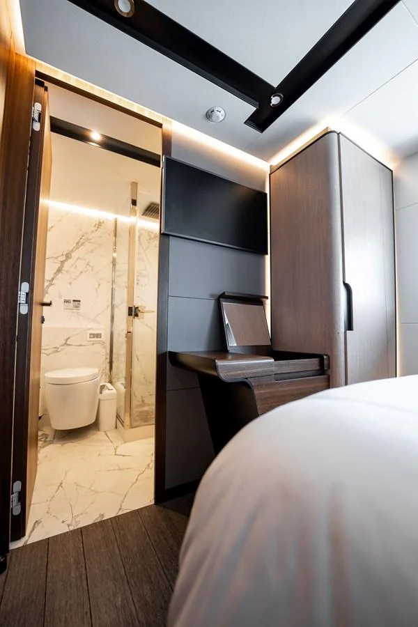 A modern stateroom featuring a sleek desk, a wall-mounted TV, and an en-suite bathroom with marble accents. aboard HOTEL COLLECTION Yacht for Sale