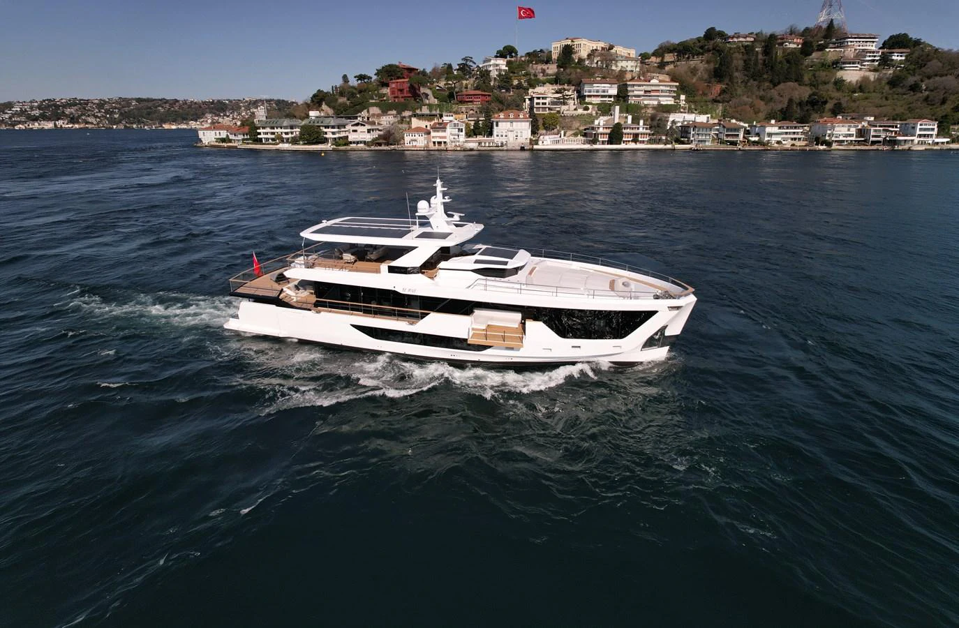 A sleek, modern yacht cruises along a scenic coastline with a backdrop of luxurious homes and greenery. aboard NUMARINE 30XP#3 Yacht for Sale