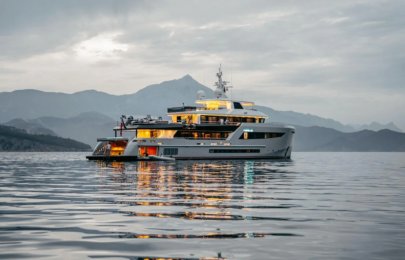 A luxurious yacht is anchored on calm waters against a backdrop of serene mountains at dusk. aboard BERING 145 Yacht for Sale