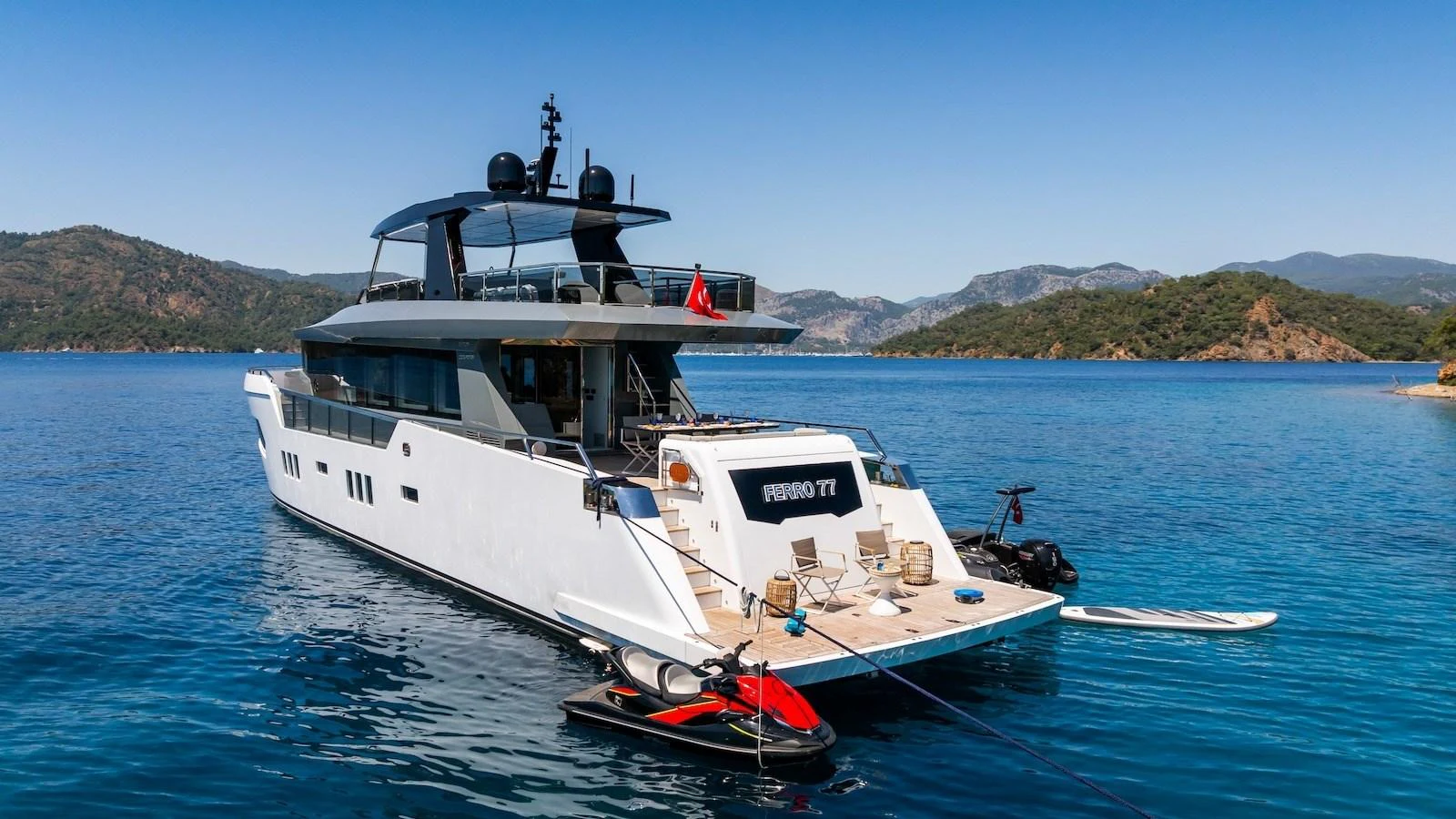 The yacht is anchored in a serene bay, showcasing its sleek aft deck and water toys. aboard FERRO 77 Yacht for Sale