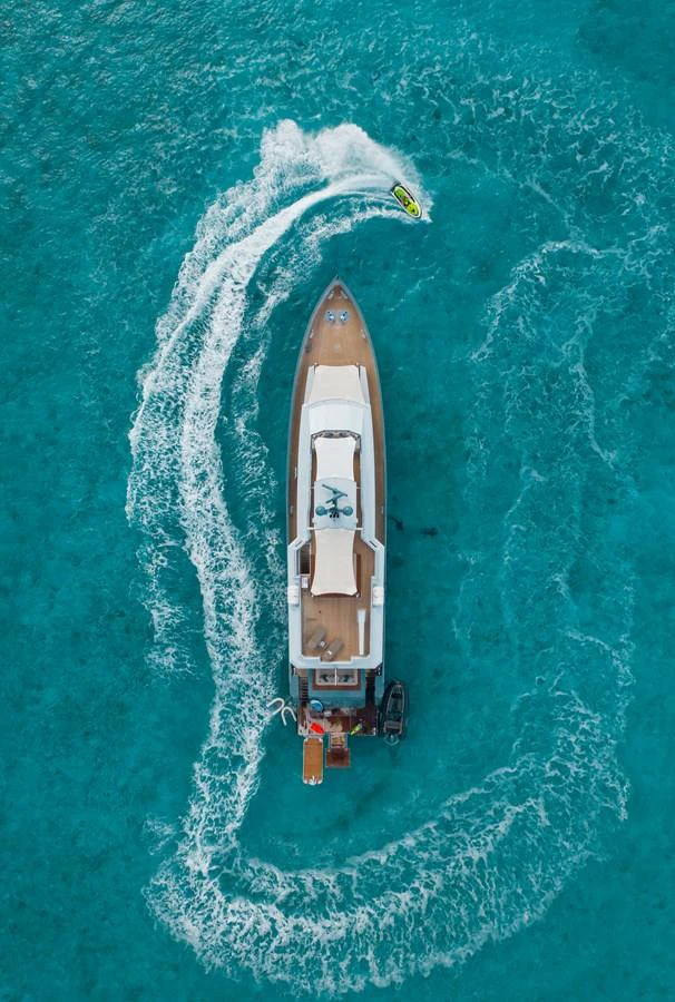 An aerial view of a yacht cruising through clear turquoise waters with a jet ski circling around it. aboard ONLY NOW Yacht for Sale