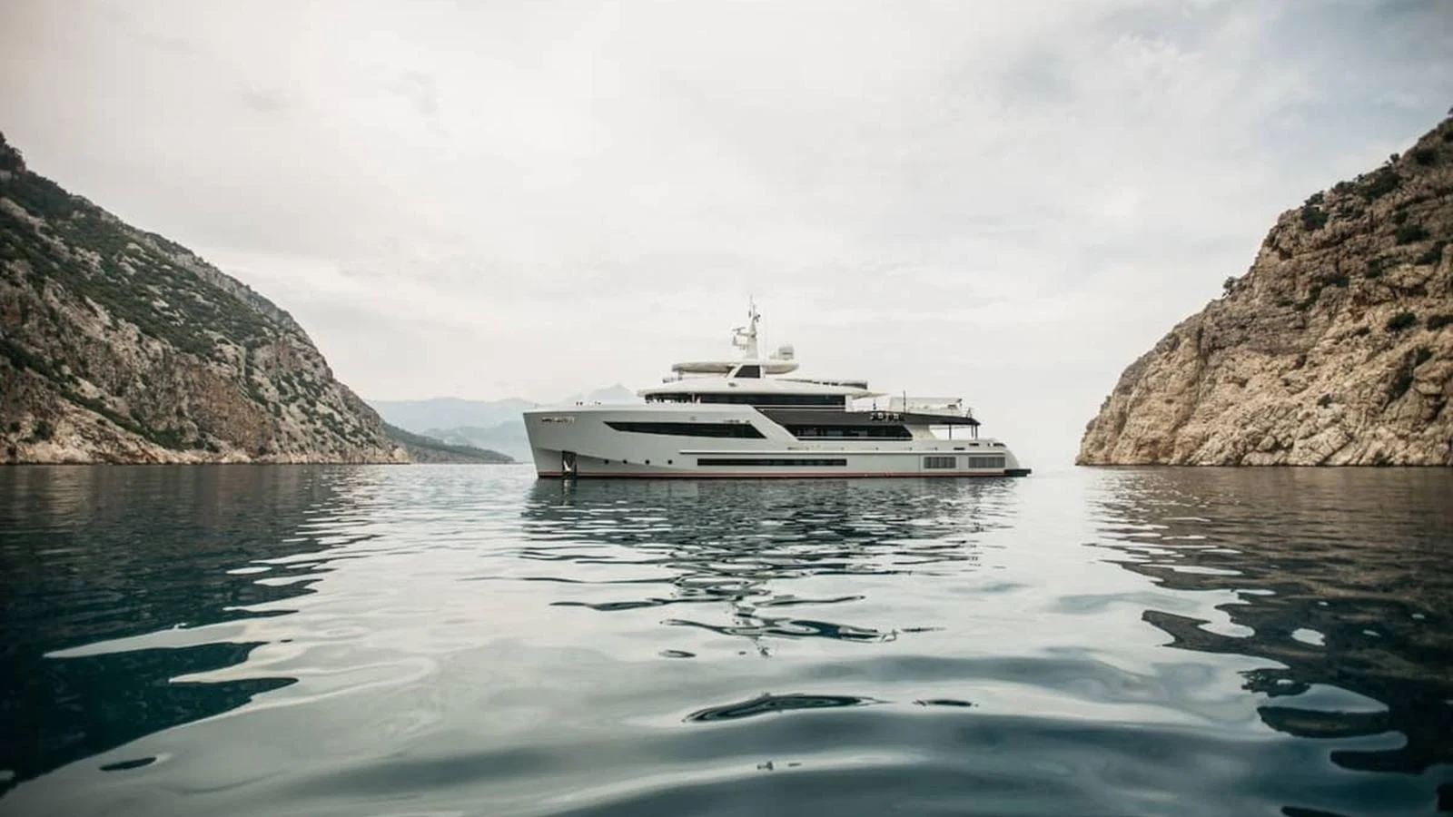 A sleek and modern yacht is anchored in a serene bay surrounded by rocky cliffs. aboard HEEUS Yacht for Sale
