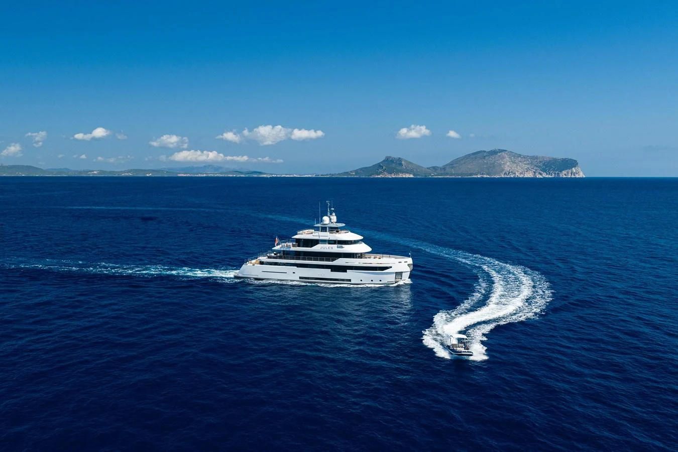 A luxurious yacht cruises through the deep blue sea with a stunning island backdrop. aboard JULES Yacht for Sale