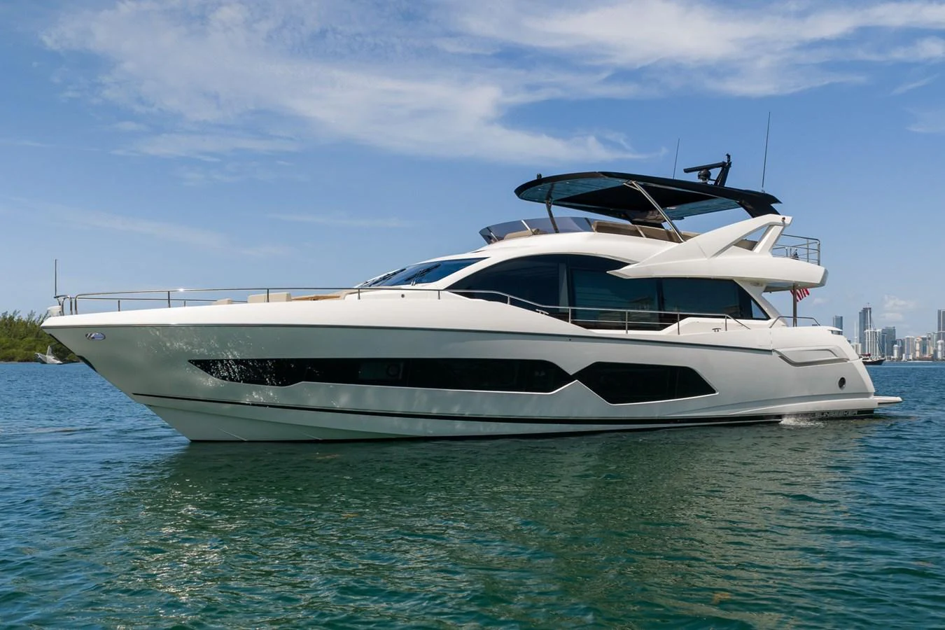 A sleek and modern yacht cruising on calm waters with a cityscape in the background. aboard BAZINGA Yacht for Sale