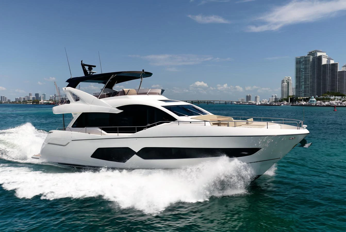 A sleek and modern yacht cruises through clear blue waters with a cityscape in the background. aboard BAZINGA Yacht for Sale