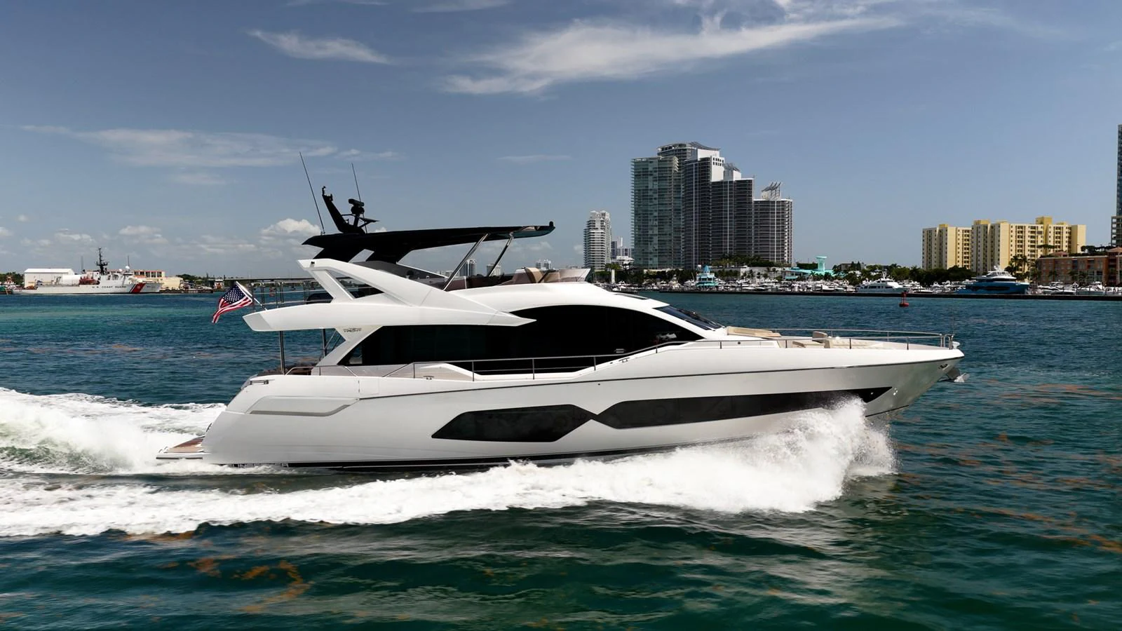 A sleek and modern yacht cruises through the water with a cityscape in the background. aboard BAZINGA Yacht for Sale