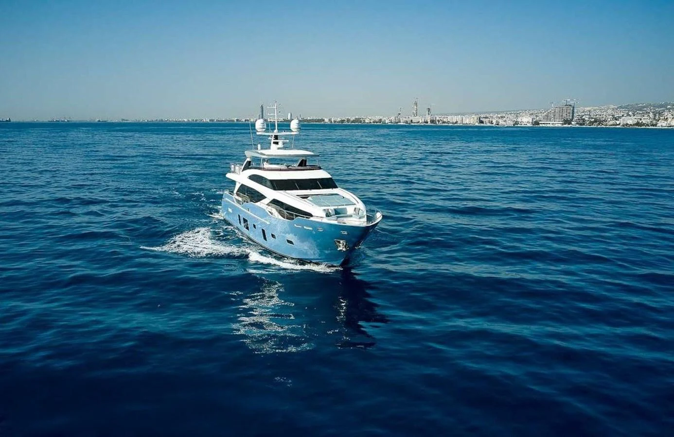 A luxurious yacht cruises through calm blue waters with a cityscape in the background. aboard ADROMELIA Yacht for Sale