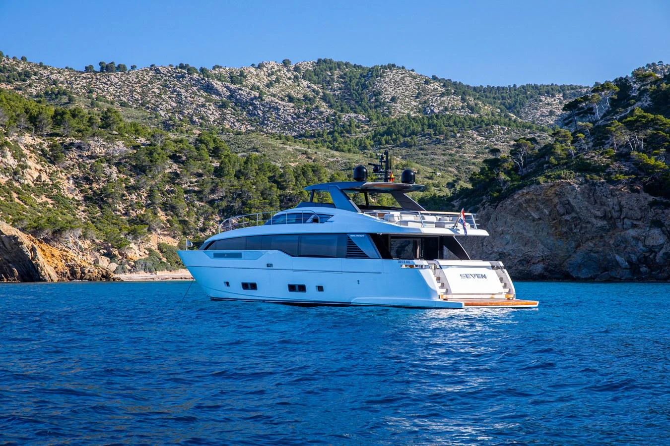 A sleek yacht is anchored in a serene bay with a mountainous backdrop. aboard SEVEN Yacht for Sale