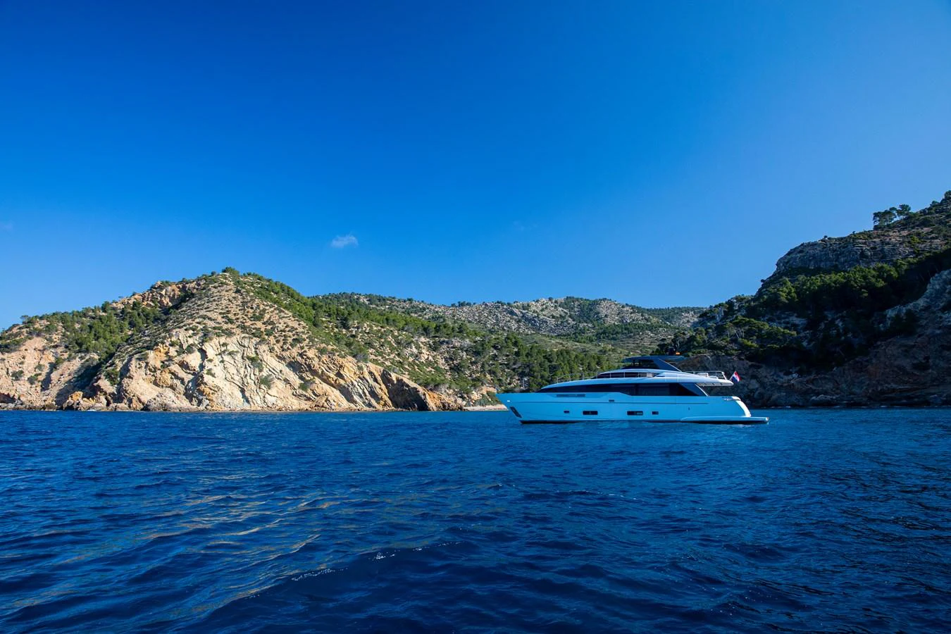 A sleek yacht cruises along a picturesque coastline under a clear blue sky. aboard SEVEN Yacht for Sale