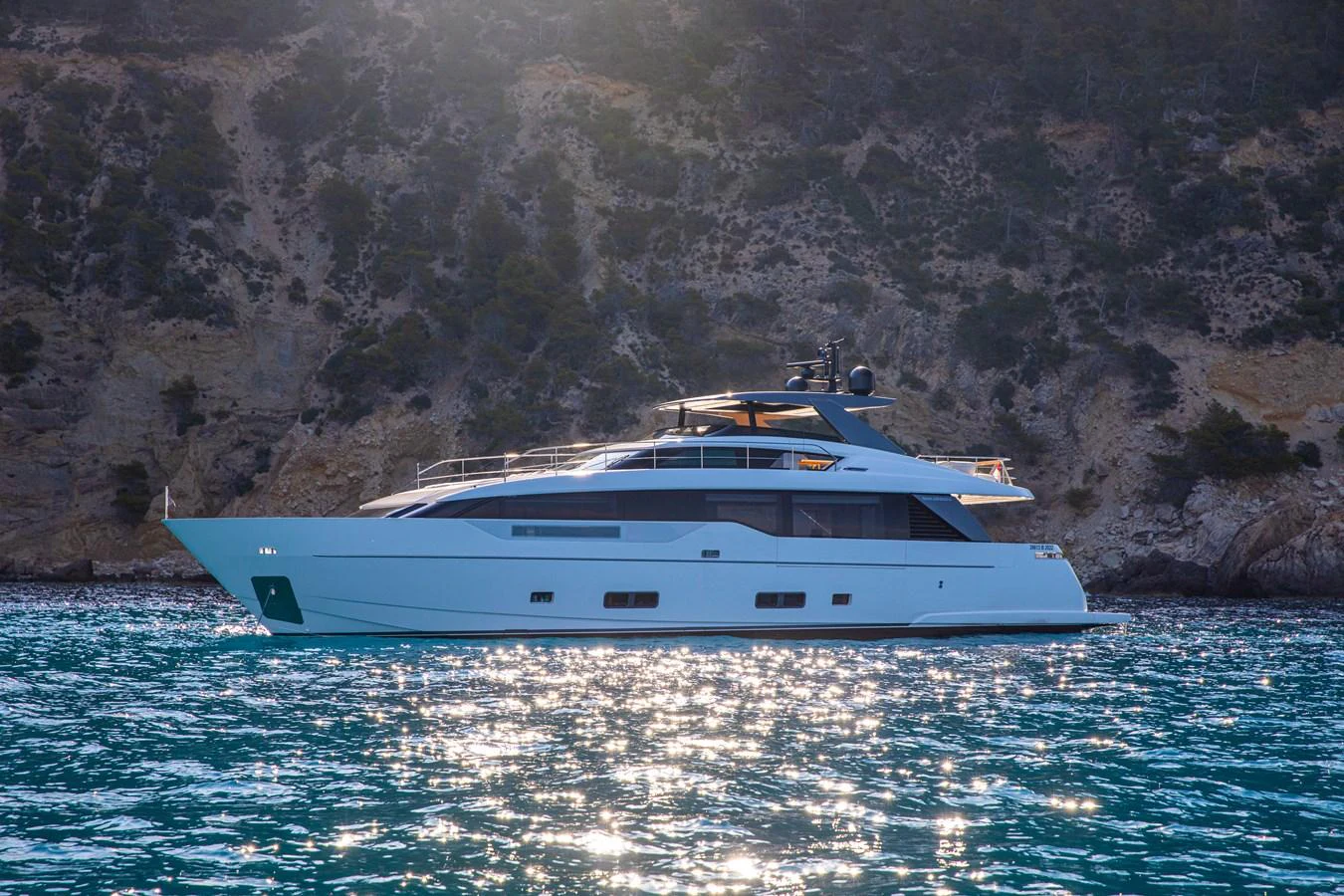 A sleek and modern yacht glides gracefully on the shimmering water against a rocky coastal backdrop. aboard SEVEN Yacht for Sale