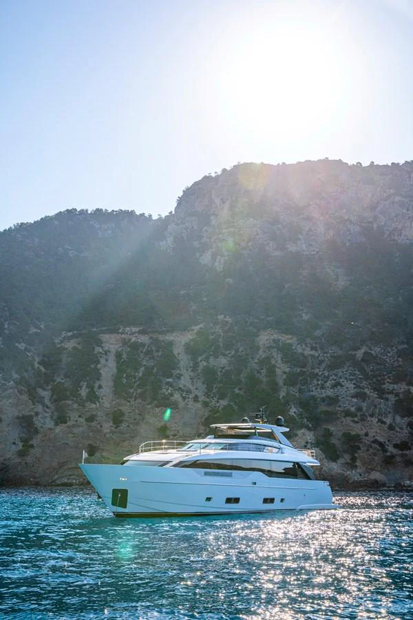A sleek yacht cruises gracefully on the water against a backdrop of rugged hills and bright sunlight. aboard SEVEN Yacht for Sale