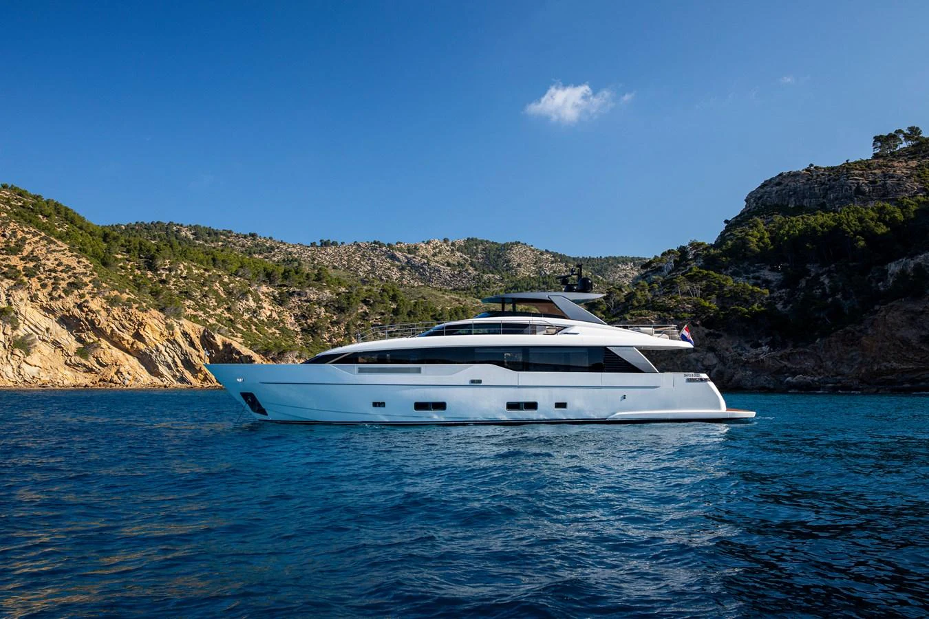 A sleek, modern yacht glides gracefully on the water against a backdrop of rugged cliffs and clear blue skies. aboard SEVEN Yacht for Sale