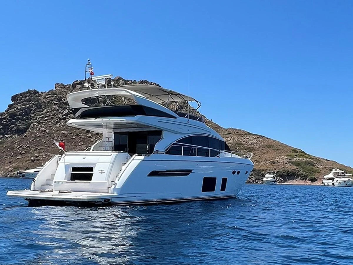 A sleek and modern yacht is anchored in a serene bay with clear blue waters and rocky hills in the background. aboard DAPHNE Yacht for Sale