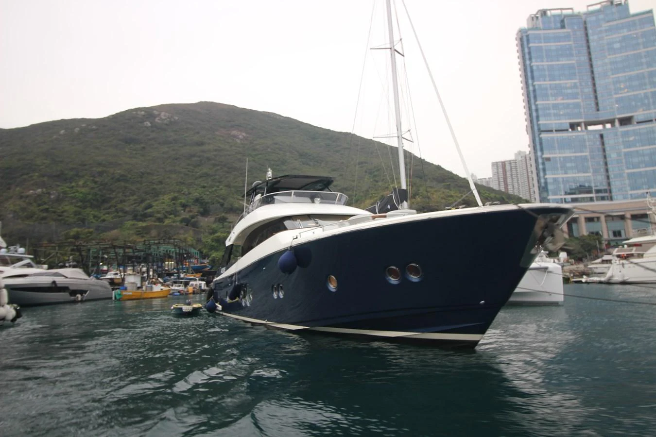 A sleek yacht is docked in a serene marina with a backdrop of lush hills and modern buildings. aboard AURORA Yacht for Sale