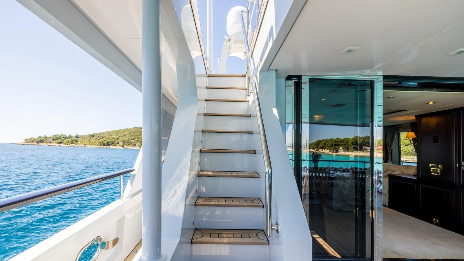 The aft deck features a staircase leading to the upper deck, with a view of the serene waters and nearby shoreline. aboard LA STELLA DEI MARI Yacht for Sale