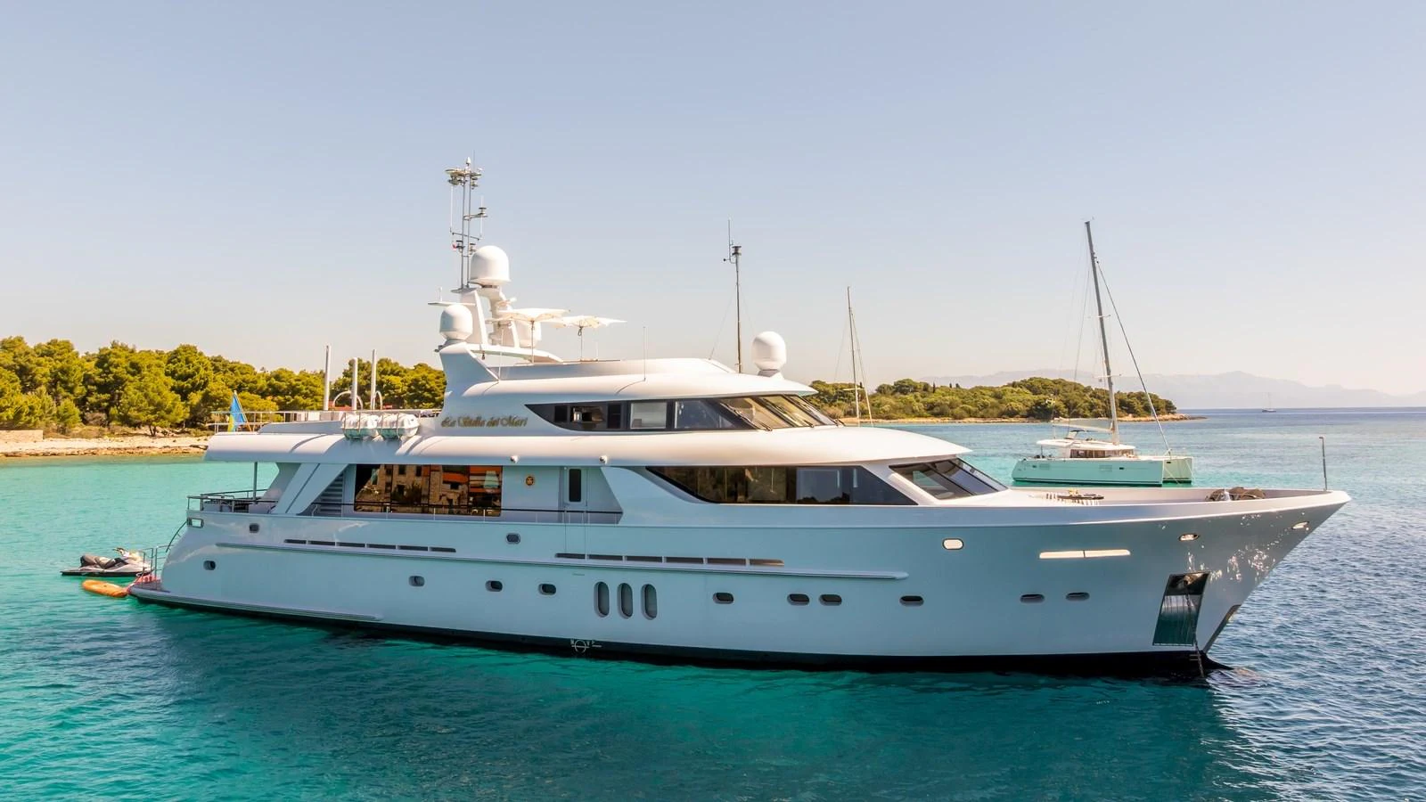 A sleek and elegant yacht is anchored in a serene, turquoise bay with a clear sky overhead. aboard LA STELLA DEI MARI Yacht for Sale