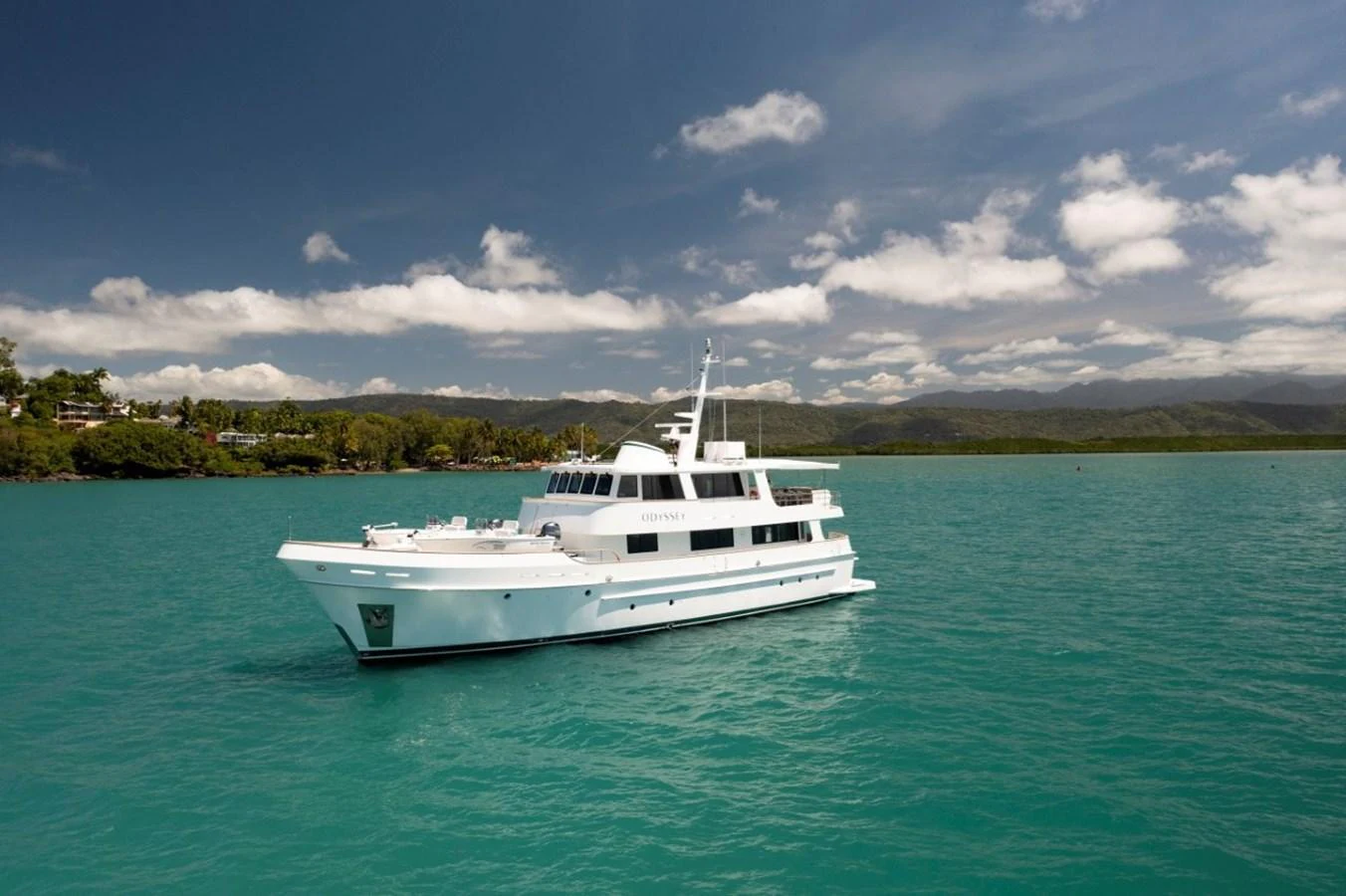 A pristine white yacht floats serenely on turquoise waters against a backdrop of lush greenery and distant mountains. aboard ODYSSEY Yacht for Sale