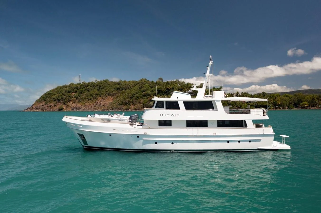 A pristine white yacht cruises on turquoise waters with a lush green island in the background. aboard ODYSSEY Yacht for Sale