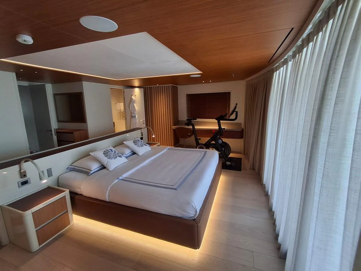 A luxurious stateroom featuring a large bed, modern decor, and an exercise bike by the window. aboard ATALI Yacht for Sale