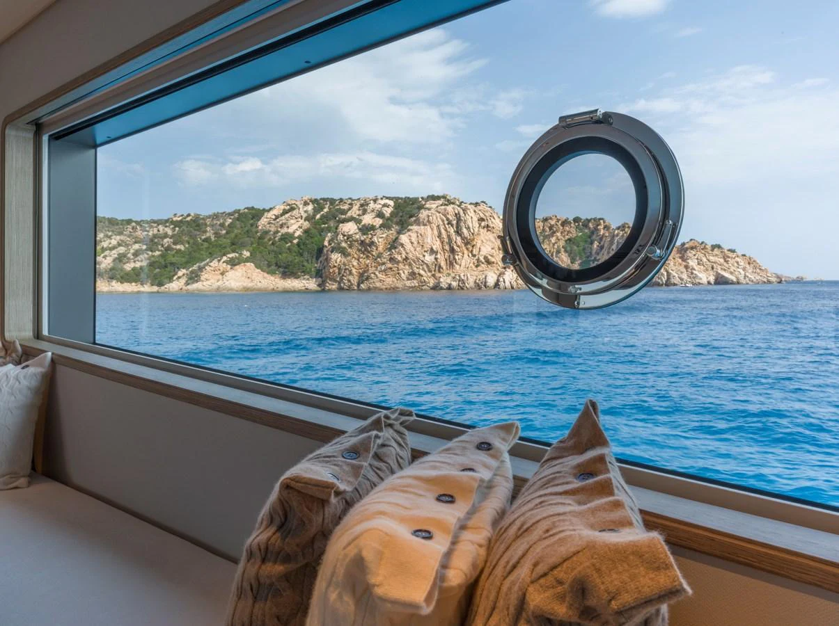 A serene view of the sea and rocky coastline through a large window with a porthole, framed by cozy pillows. aboard CLAIRE DE LUNE Yacht for Sale