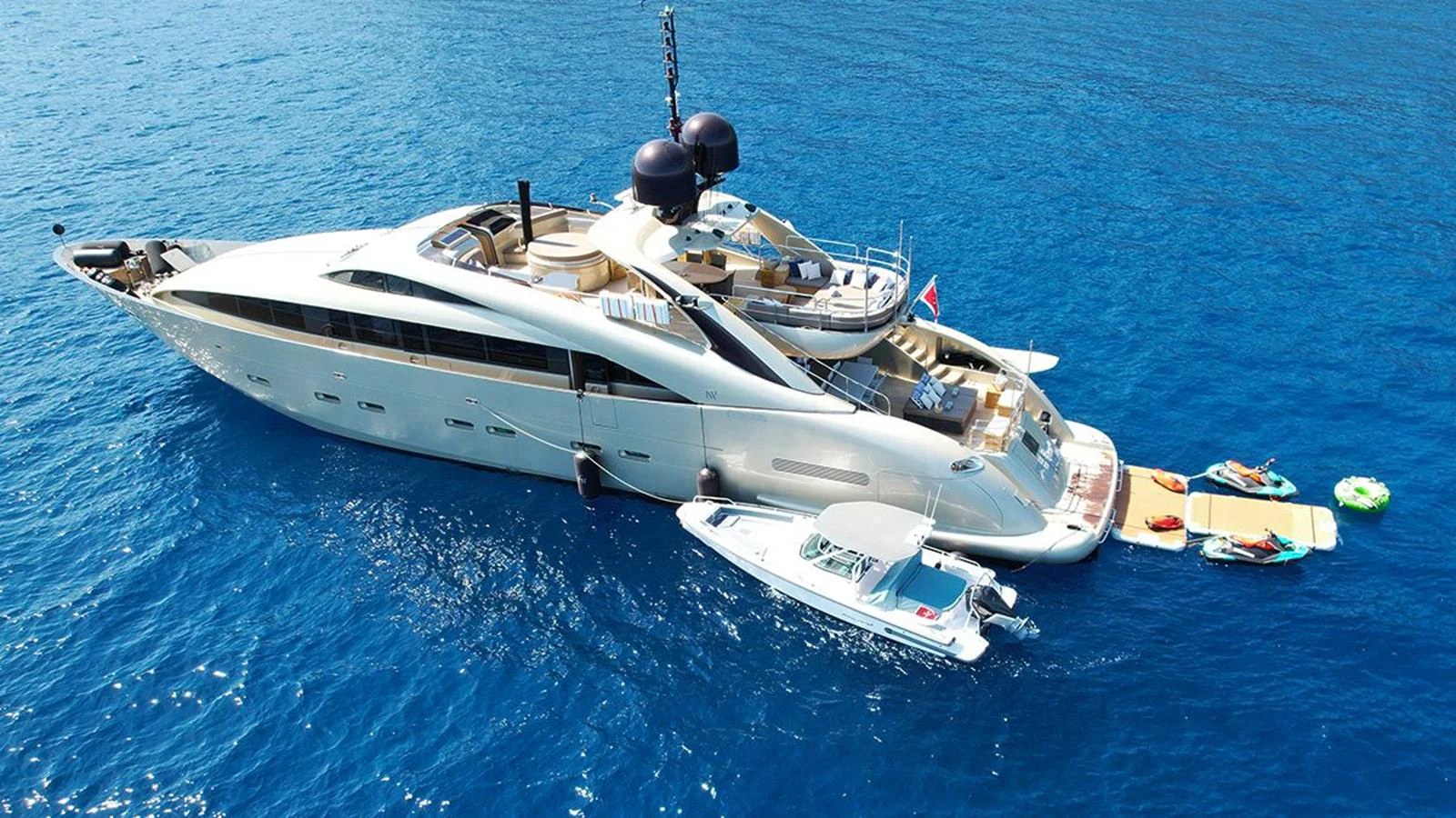 A luxurious yacht anchored in clear blue waters, accompanied by various water toys and a tender. aboard YCM 120 Yacht for Sale