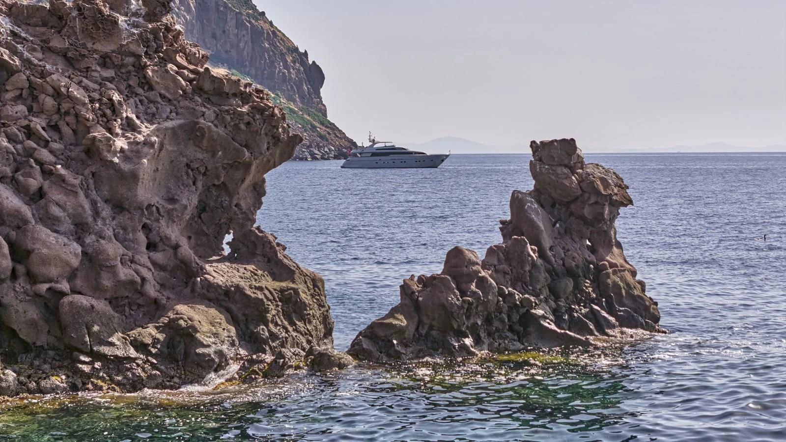 A sleek yacht cruises near rocky coastal cliffs under a clear sky. aboard OASIS Yacht for Sale