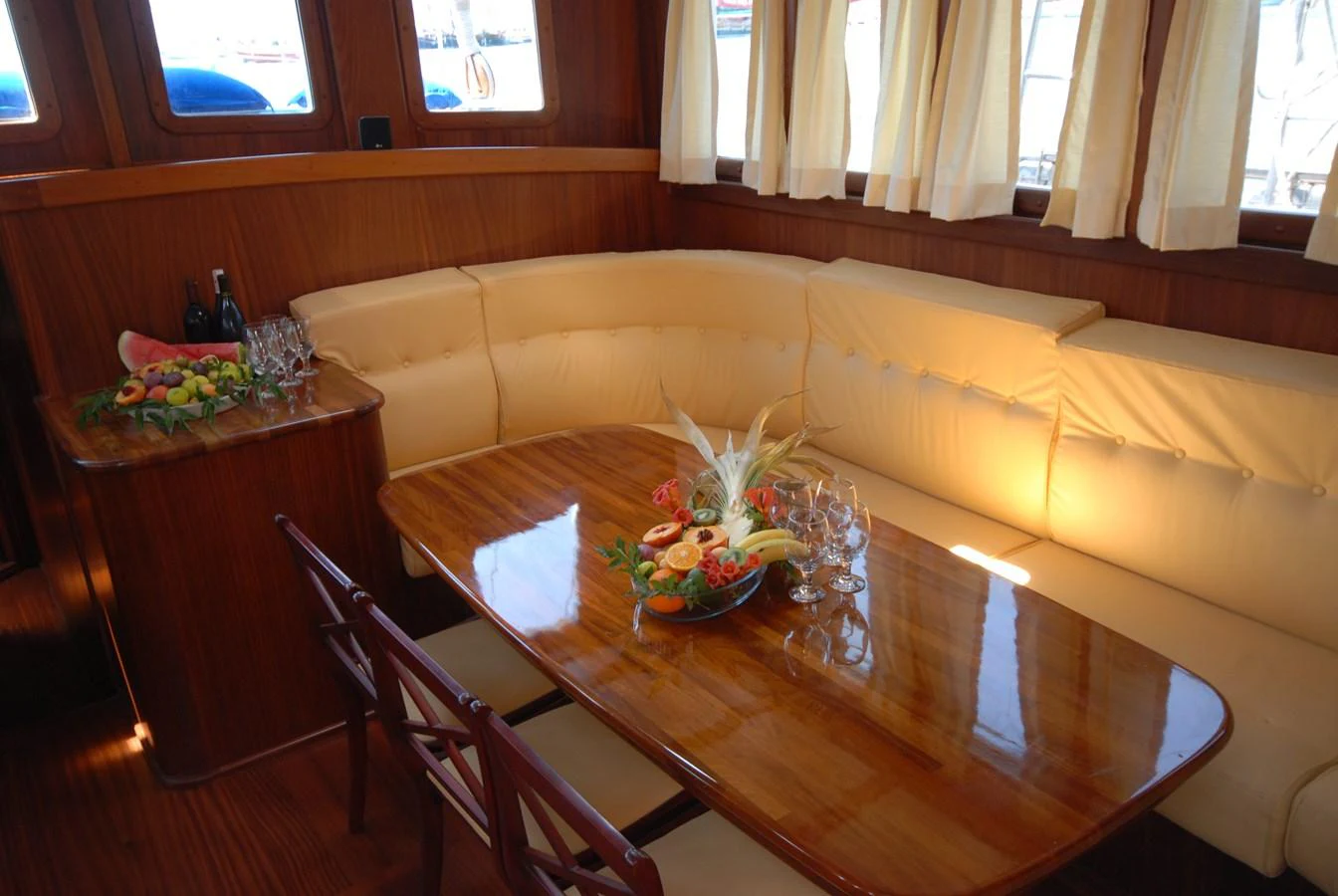The yacht's dining area features a polished wooden table surrounded by a comfortable cream-colored sofa, with a fruit platter and wine set for an elegant meal. aboard ARINNA Yacht for Sale