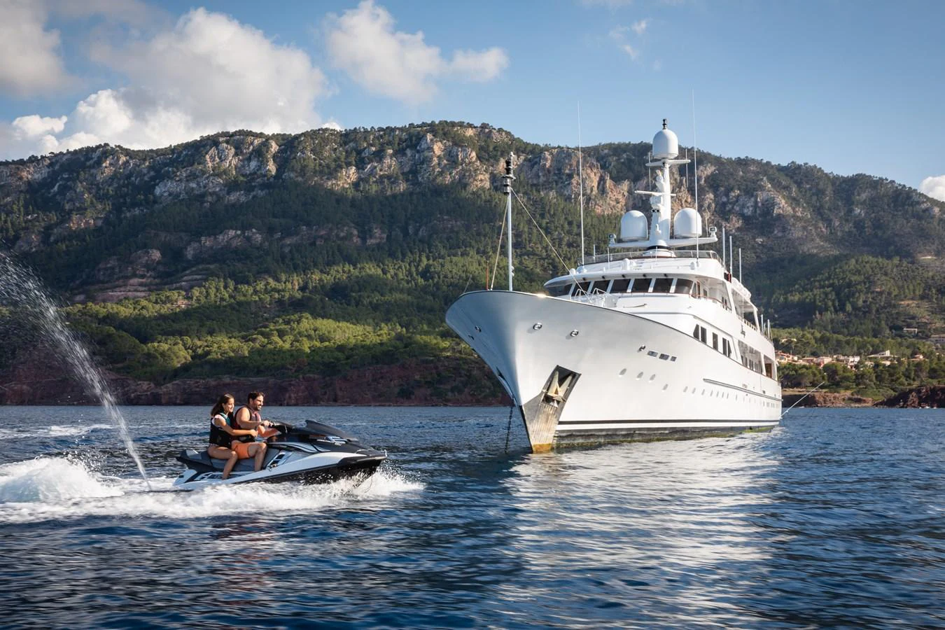 A luxurious yacht is anchored in a scenic bay, with a jet ski in the foreground. aboard MIRAGE Yacht for Sale