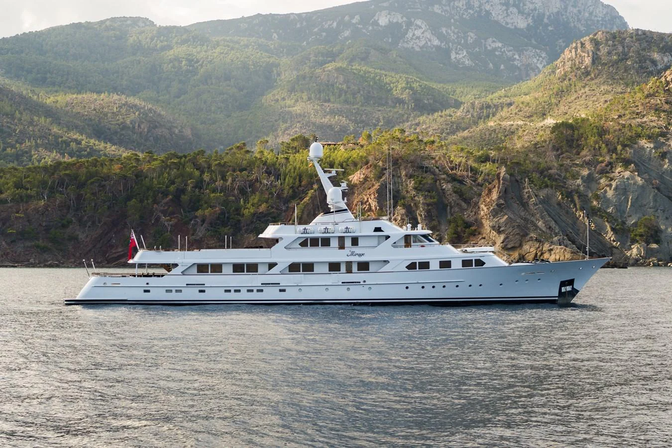 A sleek yacht cruises along a scenic coastline with mountainous terrain in the background. aboard MIRAGE Yacht for Sale