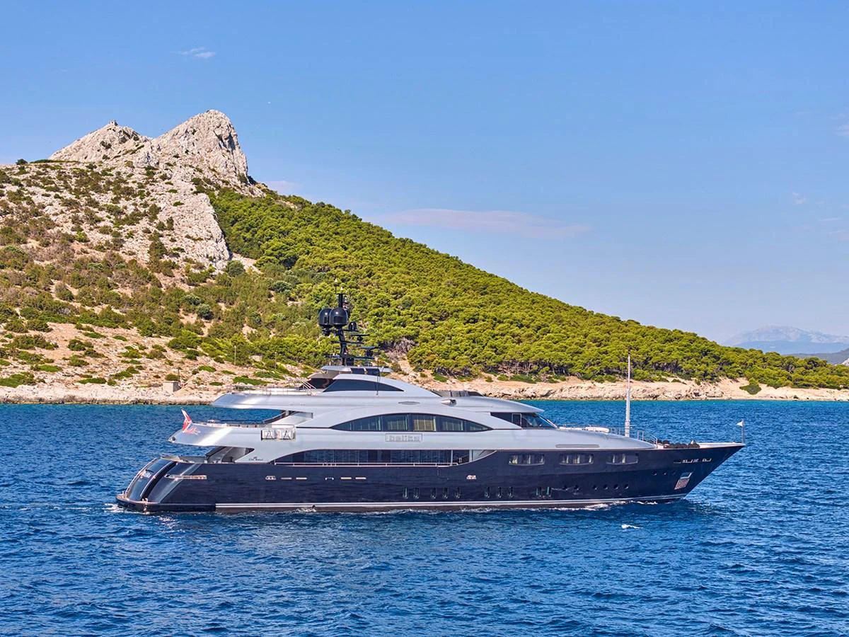 A sleek and modern yacht cruises along a scenic coastline with lush greenery and rocky hills. aboard BELITA Yacht for Sale