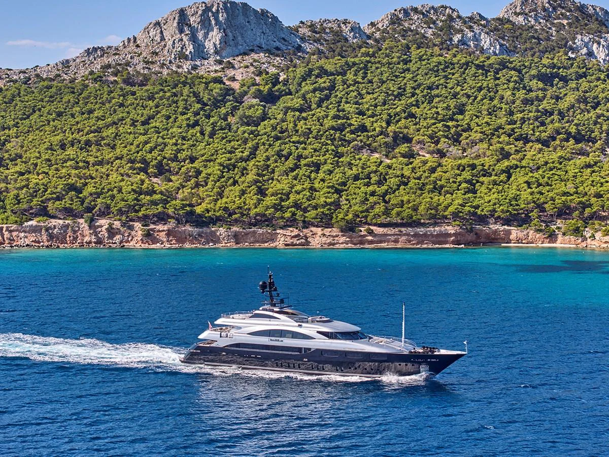 A sleek yacht cruises along a stunning coastline with clear blue waters and lush greenery. aboard BELITA Yacht for Sale