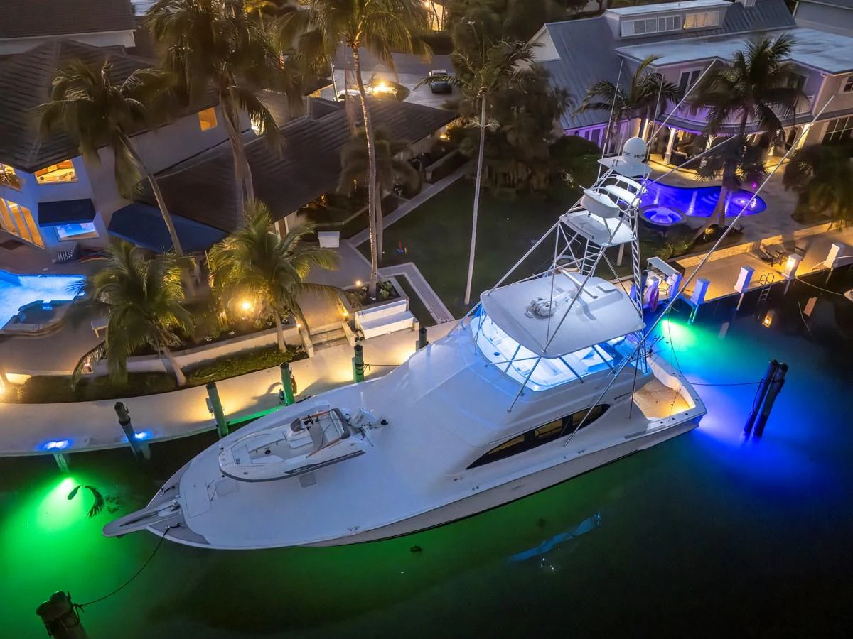 A sleek yacht is docked at a marina, illuminated by vibrant underwater lights and surrounded by palm trees and luxury homes. aboard REEL TEXAN Yacht for Sale