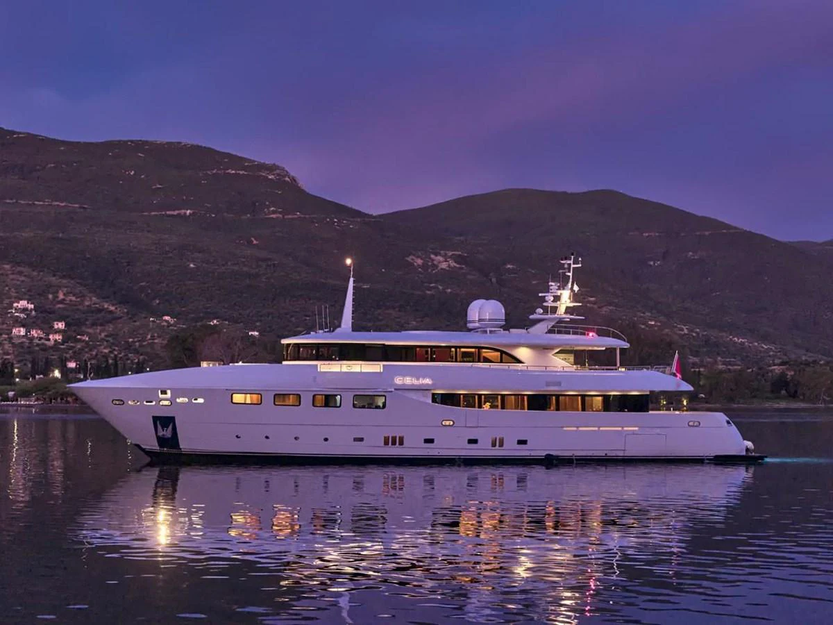 A sleek and modern yacht is gracefully anchored on calm waters at dusk, with a picturesque mountainous backdrop. aboard ULYSSES Yacht for Sale