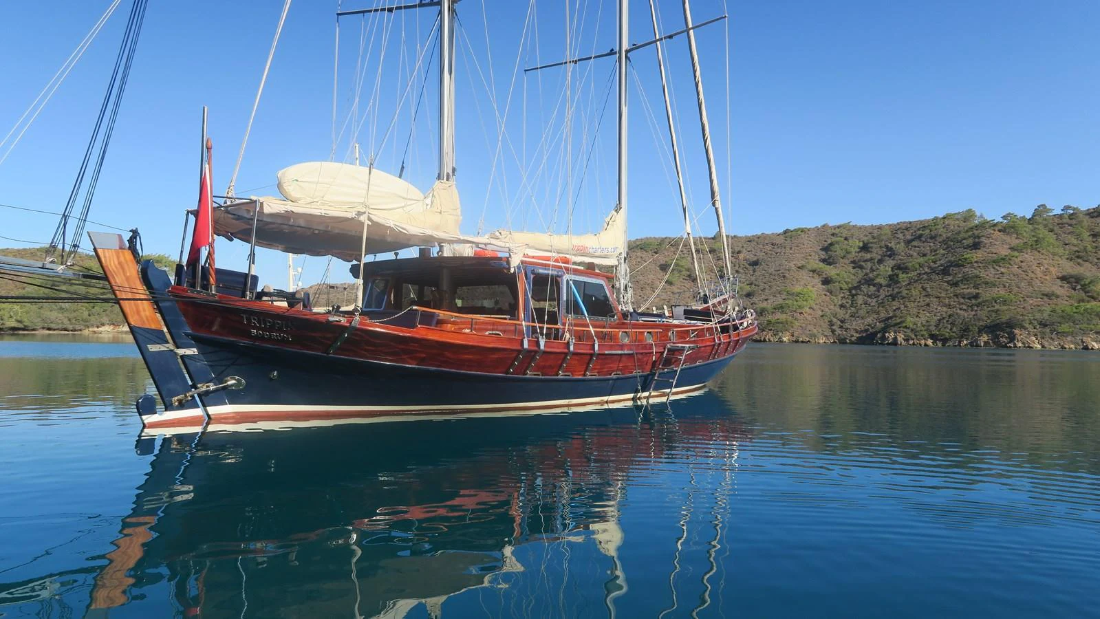 A classic wooden yacht is anchored in a serene bay with clear blue waters and a backdrop of lush hills. aboard TRIPPIN Yacht for Sale