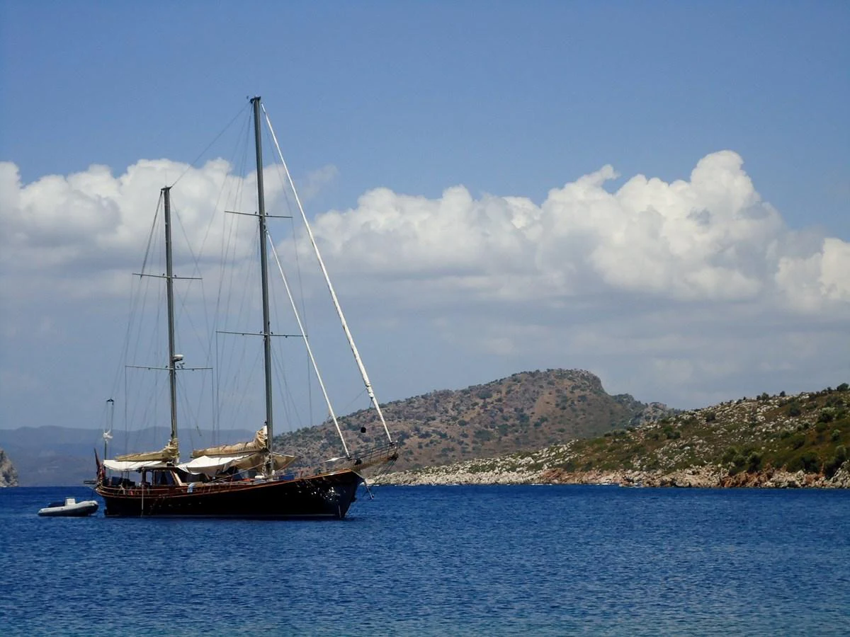 A classic sailing yacht is anchored in a serene bay with a picturesque backdrop of hills and clouds. aboard TRIPPIN Yacht for Sale