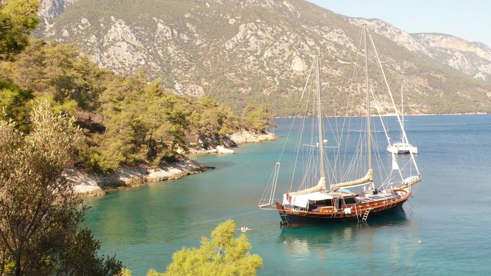 A classic yacht is anchored in a serene bay surrounded by lush greenery and mountains. aboard TRIPPIN Yacht for Sale