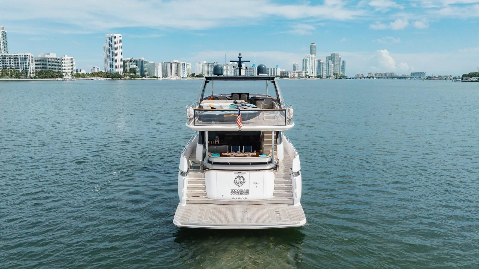 The yacht is seen from the rear, showcasing its spacious aft deck and modern design against a cityscape backdrop. aboard TOMORROW NEVER KNOWS Yacht for Sale