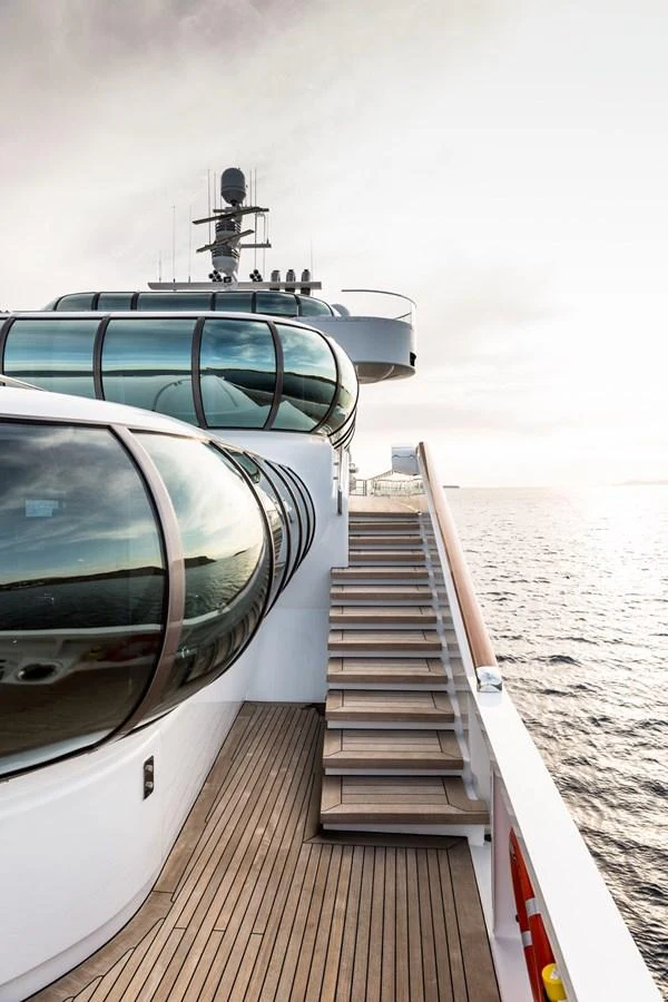 A sleek and modern yacht exterior with curved glass windows and a wooden deck overlooking the sea. aboard ZEUS Yacht for Sale