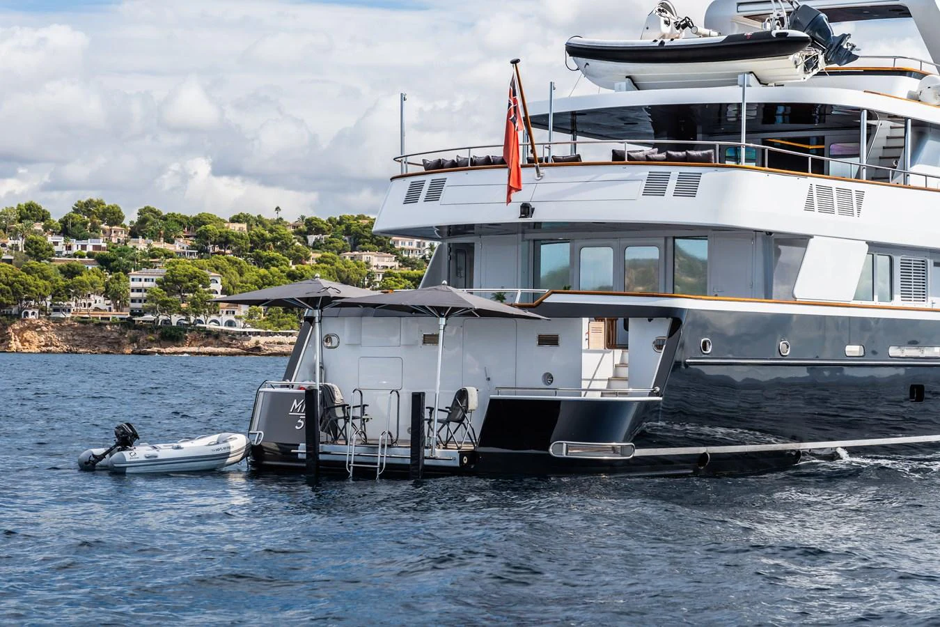 A luxurious yacht anchored near the shore, featuring a spacious aft deck with seating and a tender boat. aboard MP5 Yacht for Sale