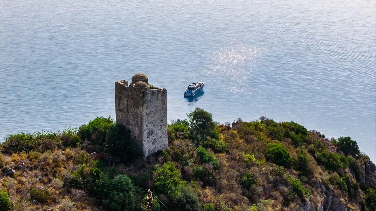 A yacht is seen cruising near a scenic coastal area with an ancient stone tower and lush greenery. aboard MAYAR Yacht for Sale