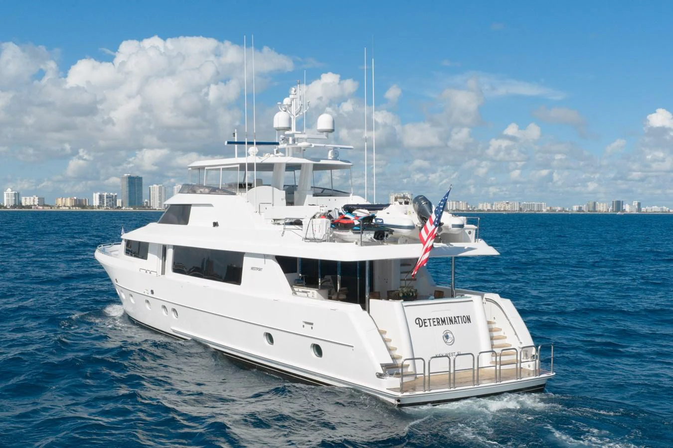 A sleek and modern yacht cruising on a clear blue sea with a cityscape in the background. aboard DETERMINATION Yacht for Sale