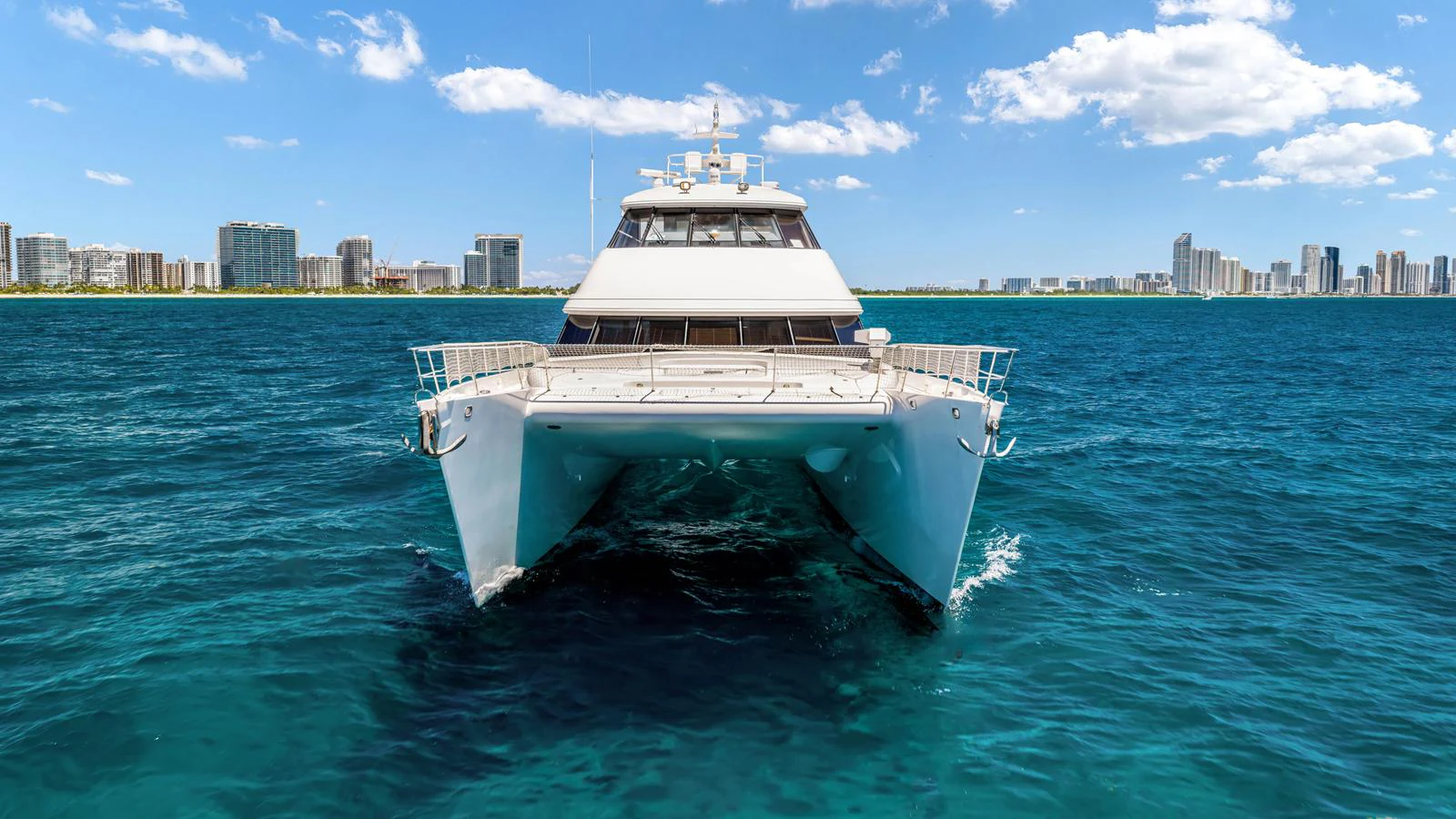A sleek catamaran yacht glides through vibrant blue waters with a city skyline in the background. aboard MOANA Yacht for Sale