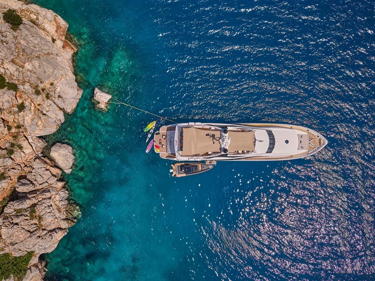 A luxurious yacht is anchored near a rocky shoreline in crystal-clear blue waters. aboard BLADE I Yacht for Sale