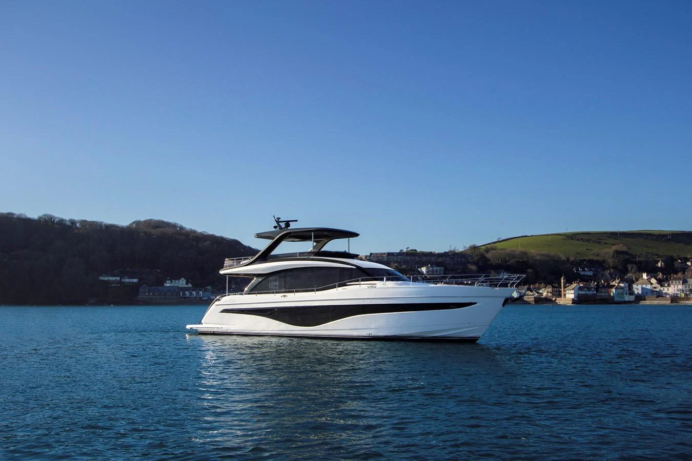A sleek, modern yacht glides gracefully on calm waters with a picturesque shoreline in the background. aboard PRINCESS Y72 Yacht for Sale
