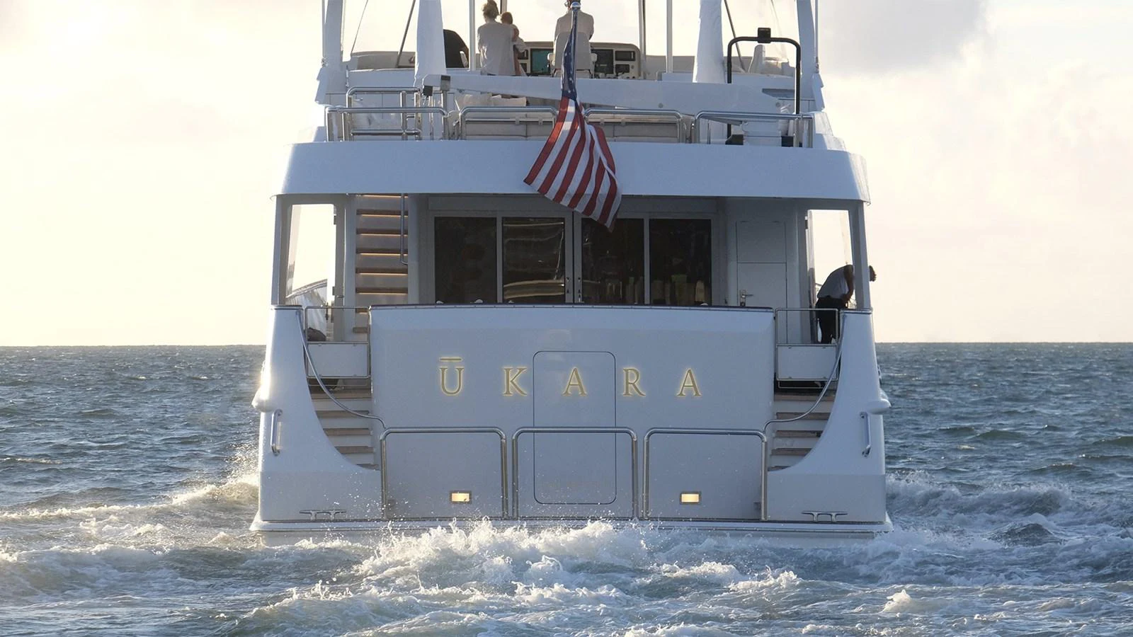 The yacht is seen from the rear, cruising on the open water with an American flag displayed prominently. aboard UKARA Yacht for Sale